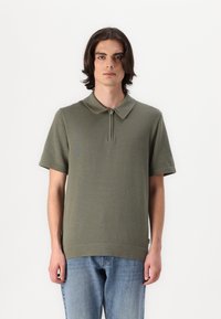 Olivgrünes Polo-Shirt mit kurzem Reißverschluss und geripptem Kragen sowie kurzen Ärmeln. Trägt man mit hellblauen Jeans aus Denim, die eine lockere Passform haben.