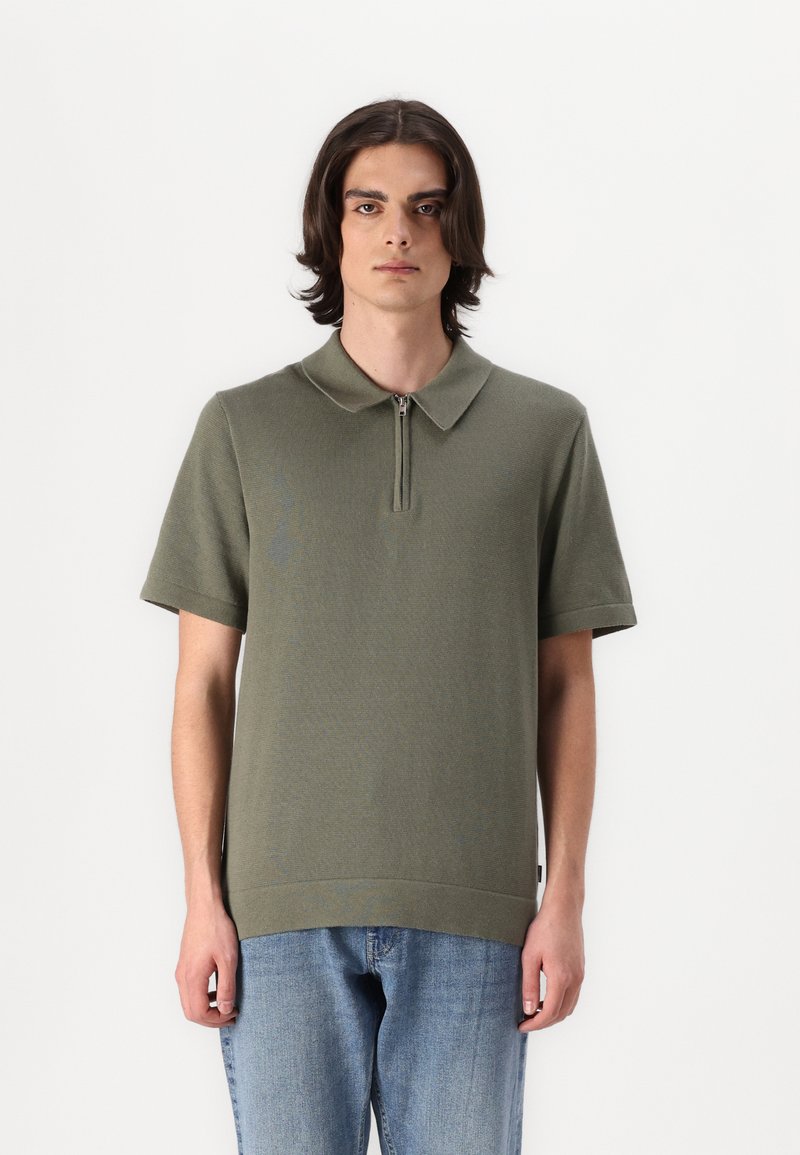 Olivgrünes Polo-Shirt mit kurzem Reißverschluss und geripptem Kragen sowie kurzen Ärmeln. Trägt man mit hellblauen Jeans aus Denim, die eine lockere Passform haben.