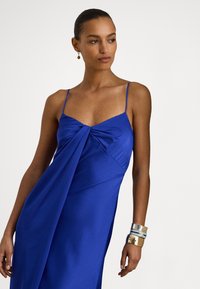 Femeie purtând o rochie albastru regal fără mâneci, cu bretele subțiri, detaliu drapat în față, brățări aurii și albastre la încheietura mâinii drepte și cercei mici rotunzi.