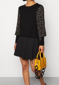 Maglione nero con maniche trasparenti a pois dorati, abbinato a una gonna svasata nera. La modella tiene una borsa gialla con una tracolla nera.