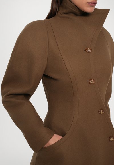 Donna che indossa un cappotto marrone aderente con colletto alto e bottoni in rilievo, con una mano nella tasca del cappotto.