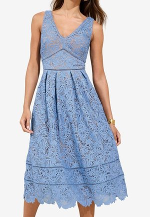 Robe midi sans manches en dentelle bleue avec un décolleté en V, taille ajustée et jupe évasée portée par une femme aux longs cheveux bruns et un bracelet doré au poignet.