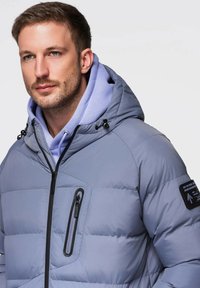 Szara pikowana kurtka z kapturem, z ukośną kieszenią na piersi zapinaną na zamek, czarnymi okuciami i teksturowanym wykończeniem.