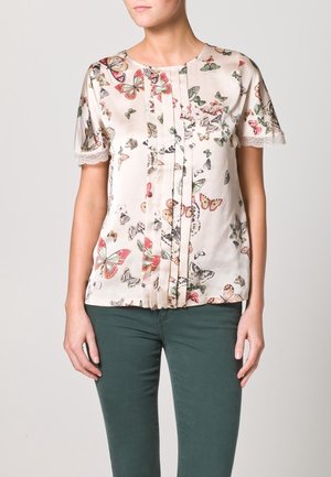 Mujer con una blusa de manga corta con estampado de mariposas multicolores y ribete de encaje en las mangas, combinada con pantalones verde oscuro.