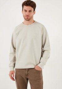 Felpa grigia chiara con maniche a raglan, colletto rotondo e un sottile logo sul petto. Abbinata a pantaloni marroni, con texture liscia.