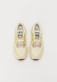 Beige Netz-Sneaker mit hellgrünen Akzenten und einer gemusterten Zunge. Sie haben eine abgerundete Zehenform und beige Schnürsenkel über einer strukturierten Oberfläche.