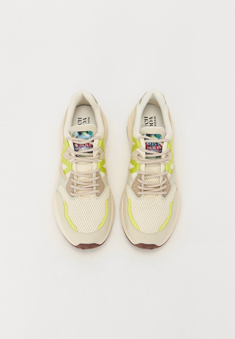 Baskets en maille beige avec des accents vert clair et une languette à motifs. Elles présentent un bout arrondi et des lacets beiges sur une surface texturée.