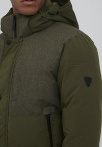 Oliven grøn jakke med en struktureret overflade. Har en hætte, sort hardware og en lynlåslomme på siden. Fremhævet med et logo-patch.