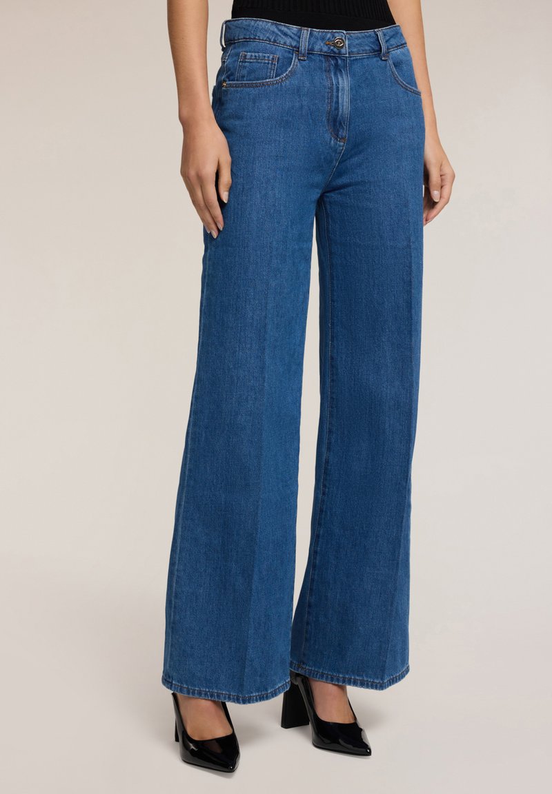 Pitkät siniset farkut leveillä lahkeilla ja korkealla vyötäröllä, joissa on etupainike, vyölenkit ja perinteiset takataskut. Tuntuva kankaantyöstö.