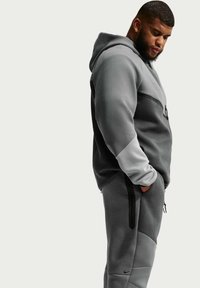 Mand i grå og sort hættetrøje og joggingbukser med hænderne i lommerne, kigger ned mod en ensartet lys baggrund.