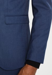 Marineblå kostymmössa med texturerad tyg. Har en enda bröstficka och en kontrasterande svart manschett. Skräddarsydd passform och strukturerad design.