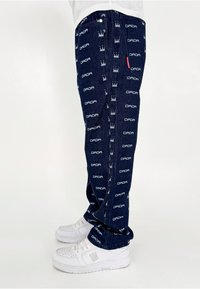 Pantalon en denim avec un fond bleu marine orné de texte "DADA" blanc répété et de motifs de couronne. Fini avec une coupe droite et des baskets blanches.