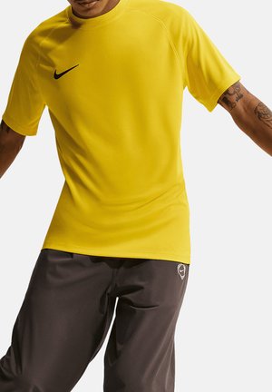 Mężczyzna ubrany w żółtą sportową koszulkę Nike z krótkim rękawem i czarne spodenki Nike, pokazany od szyi do połowy uda z widocznymi tatuażami na ramionach.