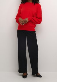 Vrouw in een rode langemouwen sweater met textuur en pofmouwen, zwarte wijde broek en zwarte hakken, staand tegen een effen muur.