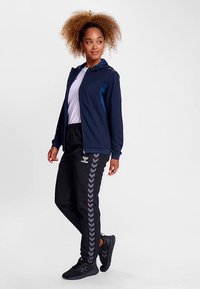 Marineblauwe hoodie met ritssluiting en lichtblauwe accenten, gecombineerd met zwarte broek met grijze chevronpatronen en zwarte sportschoenen.