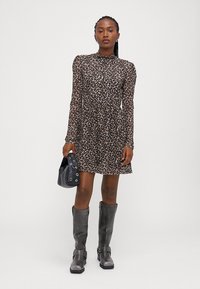 Robe longue à manches longues imprimée léopard avec un col montant, silhouette fluide, associée à des bottes noires à hauteur de genoux et un petit sac noir.
