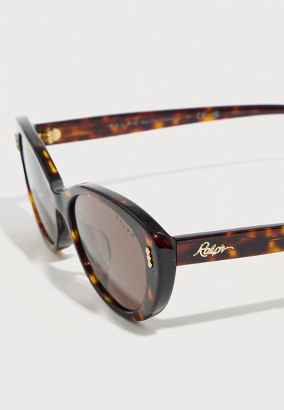 Οβάλ γυαλιά ηλίου με σχέδιο χελώνας, σκουρόχρωμοι φακοί και χρυσό λογότυπο "Ralph" στο πλάι, τοποθετημένα με κλίση σε λευκή επιφάνεια.