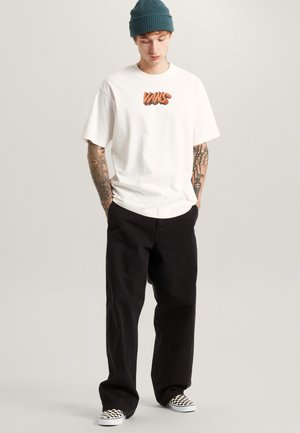 Junger Mann trägt ein weißes Vans-T-Shirt, schwarze Hose, karierte Slip-On-Schuhe, eine türkisfarbene Mütze und sichtbare Arm-Tattoos, steht mit den Händen in den Taschen.