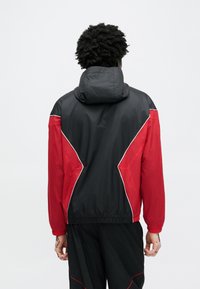 Μπουφάν αδιάβροχο σε μαύρο και κόκκινο χρώμα με κουκούλα, που διαθέτει αντιθετικά πάνελ και ελαστικά μανίκια, κατασκευασμένο από ελαφρύ, λεία υλικό.