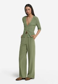 Grön stickad kofta med djup V-ringning och korta ärmar, ihop med vida gröna byxor och bruna sandals. Slät textur och avslappnad passform.