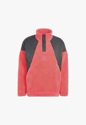 Hombre con un jersey polar coral y gris oscuro con cuello alto y pantalones negros, mirando de lado.
