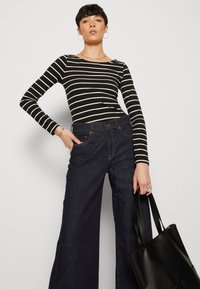 Gestreiftes schwarz-weißes Langarmoberteil kombiniert mit dunkelblauen weit geschnittenen Jeans. Hält eine schwarze Tragetasche. Glatte Stofftextur, lässige Passform.