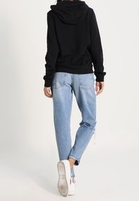 Person som bär en svart huvtröja, ljusblå slitna jeans och vita sneakers, som går bortåt mot en plain ljus bakgrund.