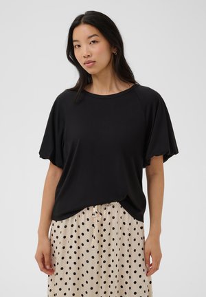 Frau mit langen dunklen Haaren trägt ein lockeres schwarzes Kurzarm-Shirt und einen cremefarbenen Rock mit schwarzen Punkten, steht vor einem schlichten Hintergrund.