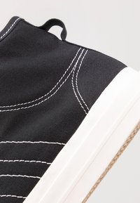 Černé plátěné vysoké tenisky s bílým prošíváním, texturovaným povrchem a kontrastní bílou gumovou podrážkou. Obsahují poutko na patě.