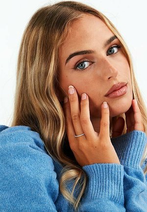 Blauer gestrickter Pullover mit ribbeliger Textur, Model mit langen welligen Haaren, dezentem Make-up und silbernem Ring an einer Hand, die nah am Gesicht ruht.