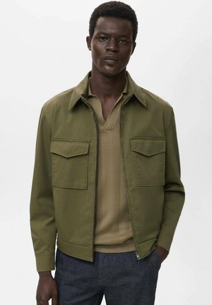 Olive green jacket with two chest pockets, zip closure, and a pointed collar, worn over a beige textured knit top. Dark trousers complete the look.