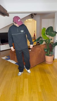 Μαύρο φλις μπουφάν με φερμουάρ και λευκές λεπτομέρειες, συνδυασμένο με φαρδιά μαύρα παντελόνια και ασημί αθλητικά παπούτσια, σε έναν μοντέρνο εσωτερικό χώρο καθιστικού.