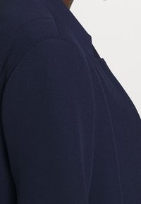 Námořnický modrý blazer s texturovanou látkou, který má strukturovaný design a ostré klopy. Detail na close-up ukazuje šev na rameni.