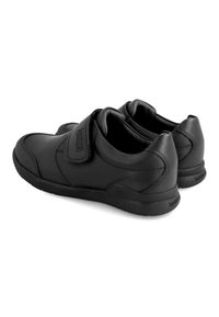 Calzado negro de cuero con forma redondeada, correa ajustable, perfil bajo y detalles de costura sutiles. Suela de goma texturizada visible.