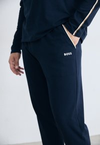 Muž v námorníckom modrom športovom oblečení s rukou v vrecku, ktorý má biele a zlaté pruhy a logo "BOSS" na nohaviciach.
