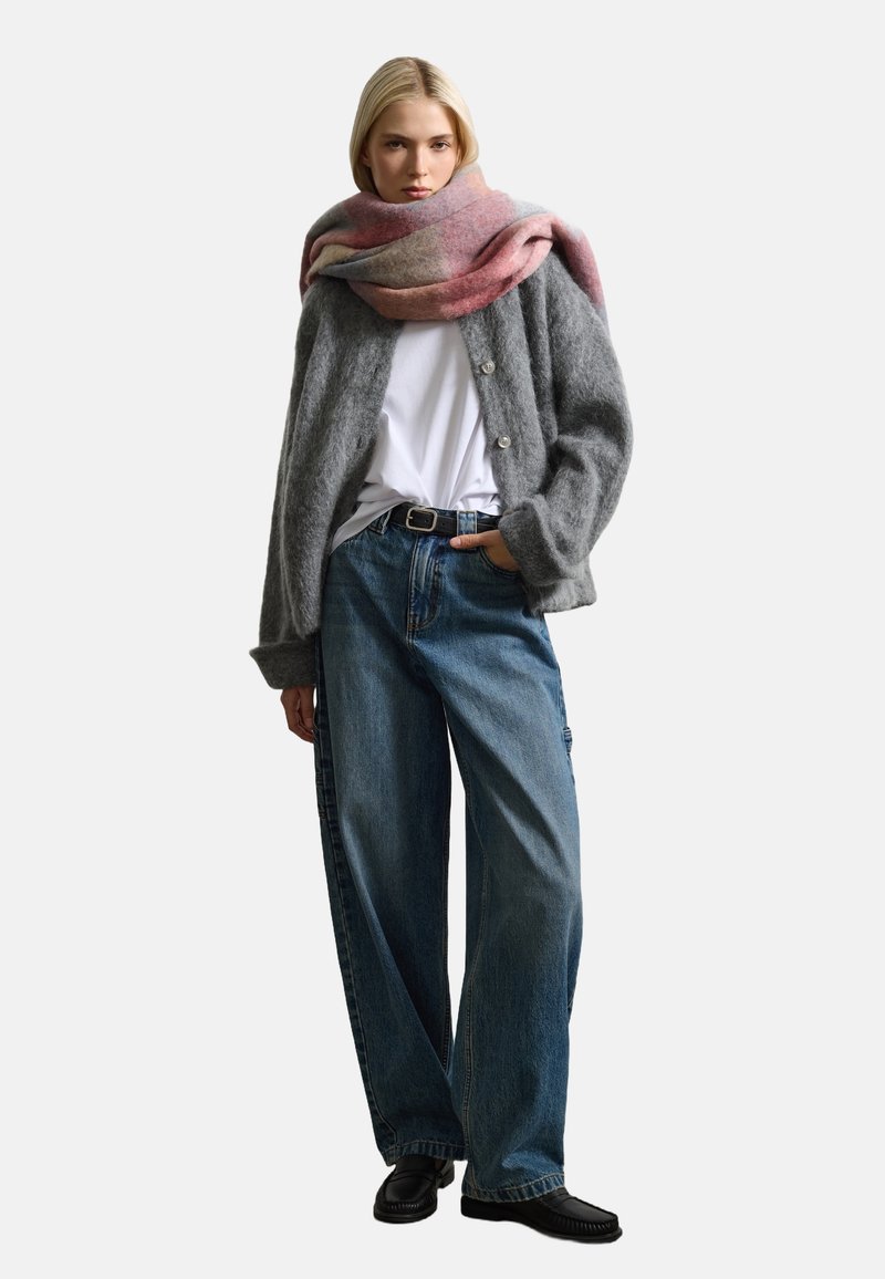 Cardigan en laine gris avec de grands boutons, t-shirt blanc, jeans en denim bleu ample et écharpe aux accents roses drapée autour du cou. Chaussures noires.