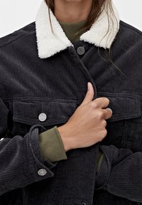 Svart kordfläcksjacka med vit konstpäls på kragen, utrustad med metallknappar och ribbade gröna ärmar som syns under ärmsluten.