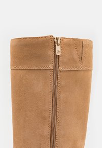 Tan suede ankle boot with a gold zipper detail along the side and decorative stitching. The top edge is slightly folded.