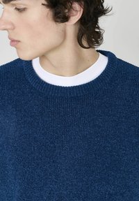 Pull en maille bleu avec un motif en relief, encolure ronde, et une chemise blanche superposée en dessous. Vue rapprochée des détails du tissu.