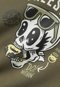 Un tissu vert présente un graphique d'un crâne de dessin animé avec des lunettes de soleil, tenant un donut avec un glaçage jaune. Comprend du texte et des icônes ludiques.