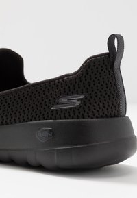 Scarpa sportiva nera con un upper in mesh testurizzato, suola in gomma spessa e un'etichetta sul retro. Presenta un logo sottile sul lato.