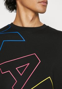 Jeune personne portant un t-shirt noir avec de grandes lettres contourées en rose, bleu et jaune, regardant légèrement sur le côté sur un fond uni.