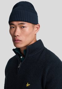 Gorro de punto acanalado en azul marino y suéter oscuro con cuello alto y cremallera de un cuarto, con un pequeño logotipo amarillo en el pecho.