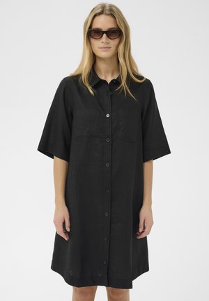 Femme aux longs cheveux blonds portant des lunettes de soleil foncées et une robe noire ample à boutons avec des manches courtes.