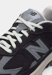 Μαύρο και γκρι παπούτσι New Balance με λευκά κορδόνια και λογότυπο στο πλάι και στη γλώσσα, εμφανιζόμενο από κοντινή γωνία.