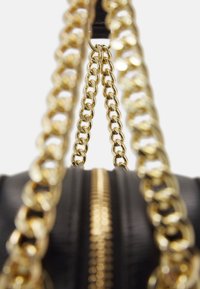 Czarna torebka z pikowaną teksturą, złotymi łańcuszkami na rączkach oraz centralnym zamkiem błyskawicznym. Widok z bliska podkreśla szczegóły łańcuszka i zamek.