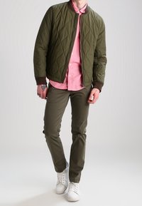 Olivfarbene, gesteppte Bomberjacke mit braunem Rippkragen, pinkem Button-up-Shirt, olivfarbenen Hosen und weißen Sneakers. Glatter Stoff und lässige Passform.