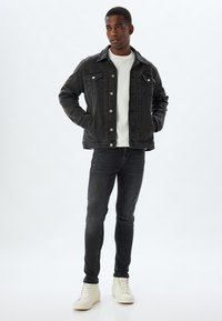 Chaqueta de mezclilla negra con cierre de botones y dos bolsillos en el pecho, combinada con jeans oscuros y zapatillas altas blancas sobre un fondo liso.