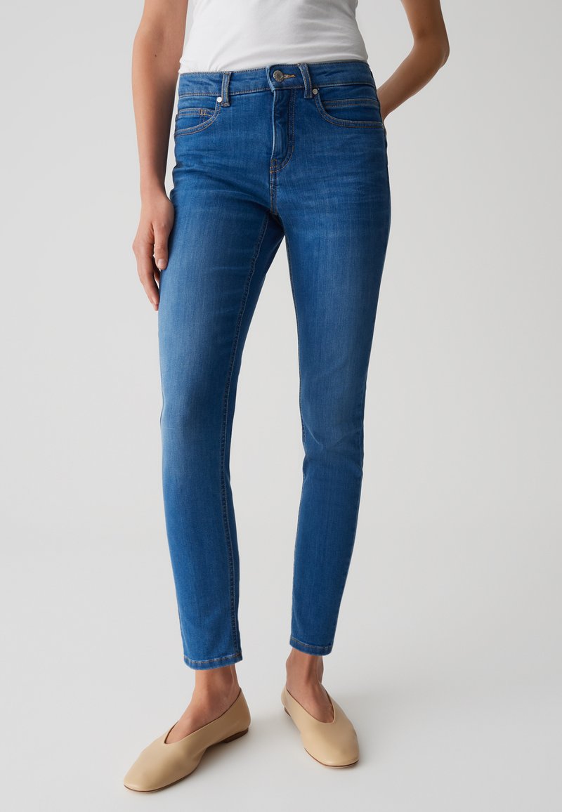 Jean skinny en denim avec décoloration légère, cinq poches et une coupe ajustée. Assortis à des chaussures plates beiges.