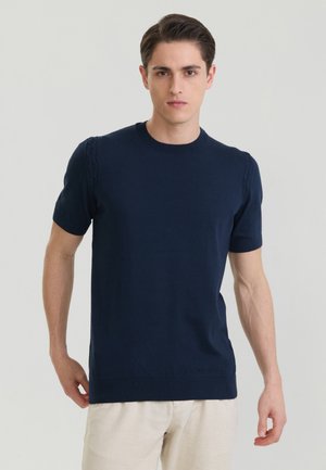 Junger Mann mit kurzen braunen Haaren, der einen marineblauen Kurzarmpullover und hellbeige Hose vor einem schlichten hellgrauen Hintergrund trägt.